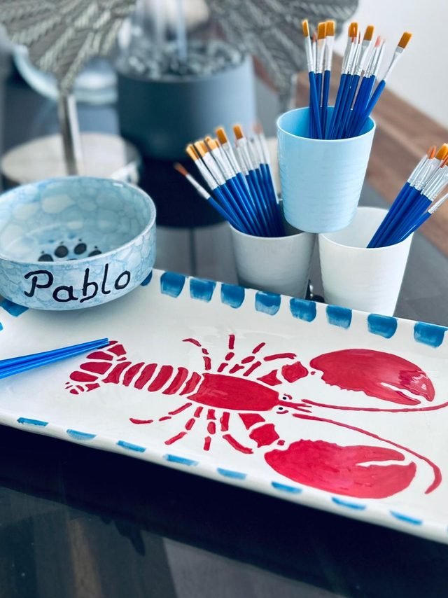 Selbst bemalte Servierplatte mit rotem Hummer und blau getupftem Rand, daneben ein Schälchen mit dem Namen Pablo und Becher voller Pinsel